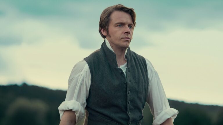 "A man in historical clothing stands outdoors with a serious expression, wearing a vest over a white shirt, with hills and trees in the blurred background under a cloudy sky."