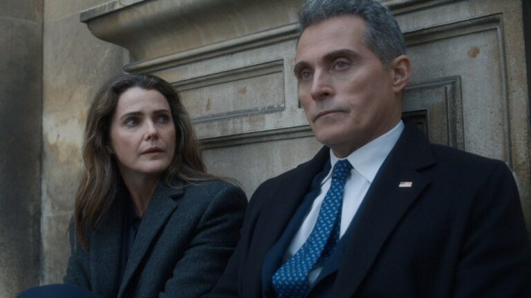 "Two people in formal coats sit on outdoor stone steps, appearing serious and deep in conversation, with an old stone wall in the background. The man wears a suit and tie with an American flag pin, suggesting a political or official context."