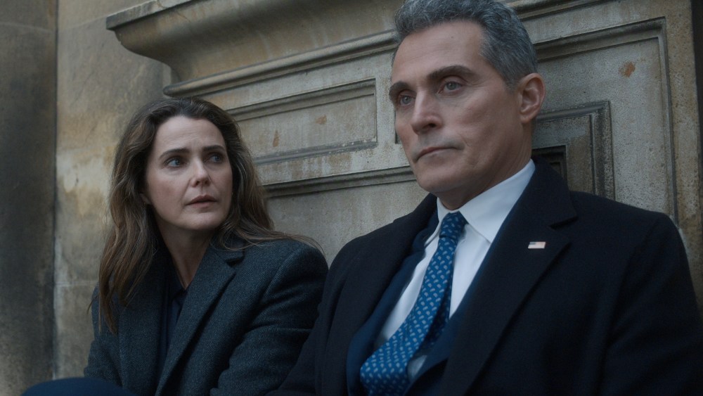 "Two people in formal coats sit on outdoor stone steps, appearing serious and deep in conversation, with an old stone wall in the background. The man wears a suit and tie with an American flag pin, suggesting a political or official context."